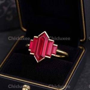 May include: A gold ring with a large, rectangular, ruby-red gemstone in a geometric design. The ring is set in a gold band and is displayed in a black jewelry box with a gold trim. The brand name "Chicluxee" is visible.