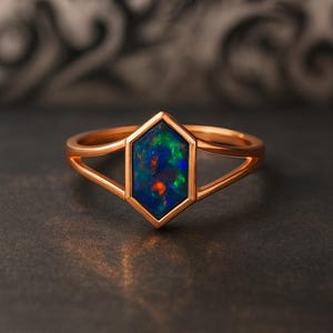 May include: A rose gold ring featuring a hexagonal opal stone with a vibrant display of blue, green, and orange colors. The ring has a delicate, open design with a band that splits into two parallel lines on either side of the stone.