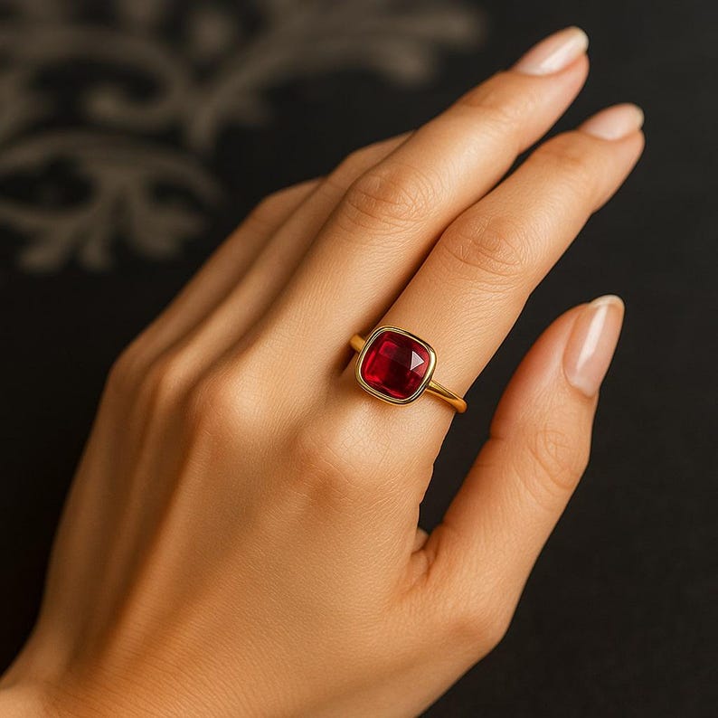 K&ouml;nnte beinhalten: Ein goldener Ring mit einem gro&szlig;en, quadratisch geschliffenen, tiefroten Edelstein. Der Ring hat ein schlichtes, elegantes Goldband. Der Edelstein ist der Blickfang und f&auml;ngt das Licht ein.
