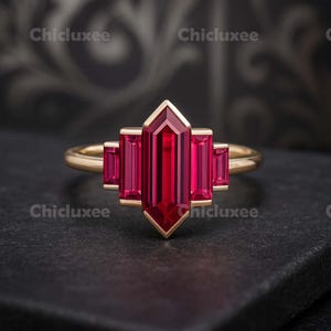 May include: A gold ring featuring a large, rectangular, deep red gemstone at the center, flanked by smaller rectangular stones. The ring has a geometric design and is set against a dark background. The word "Chicluxee" is printed in white in the corners.
