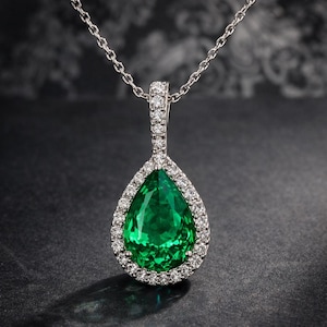Puede incluir: Un collar de plata con un colgante en forma de pera. El colgante presenta una gran gema verde vibrante rodeada por un halo de pequeñas gemas claras. La cadena es delicada y el engaste es detallado.