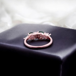May include: A rose gold ring with a floral design, featuring green gemstones. The ring is set on a dark surface, with a soft, white background. The ring's intricate details and color contrast are highlighted.