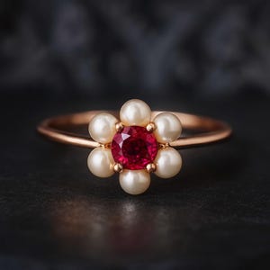 May include: A gold ring with a flower design. The centre of the flower is a round, red gemstone, encircled by six white pearls. The ring is set against a dark background.