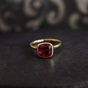 K&ouml;nnte beinhalten: Ein goldener Ring mit einem quadratischen, tiefroten Edelstein. Der Ring hat ein schlichtes, elegantes Design mit einem polierten Goldband. Der Edelstein ist in einer L&uuml;nettenfassung gefasst, was seine satte Farbe und Brillanz unterstreicht.
