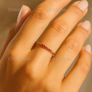 May include: A gold ring with a row of small, vibrant red gemstones set across the top. The ring is thin and delicate, with a polished finish. The ring is shown on a person's finger.