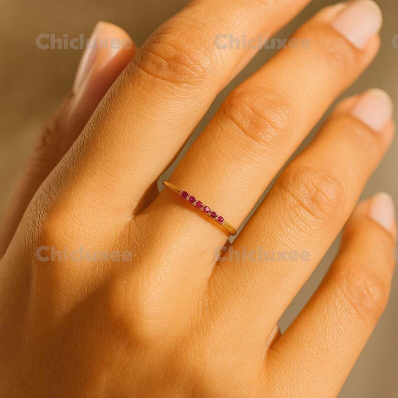 May include: A gold ring with a row of small, vibrant red gemstones set across the top. The ring is thin and delicate, with a polished finish. The ring is shown on a person's finger.
