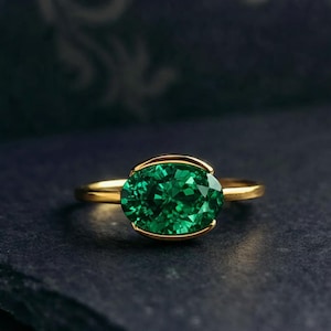 May include: A gold ring featuring a large, oval-cut emerald gemstone. The emerald is a vibrant green color and is set in a gold bezel. The ring has a simple, elegant design.
