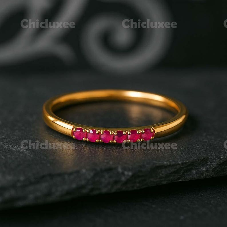 May include: A gold ring with a row of small, vibrant red gemstones set across the top. The ring is displayed on a dark, textured surface, highlighting its polished finish and the brilliance of the stones.