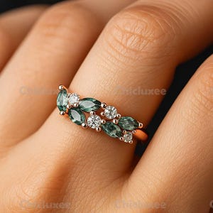May include: A rose gold ring featuring a leaf-like design with alternating green and white gemstones. The ring's design incorporates marquise-cut green stones and round, clear stones, creating a nature-inspired aesthetic.