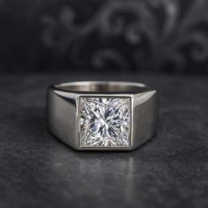 Op de afbeelding: Een zilveren ring met een vierkant geslepen, heldere edelsteen. De ring heeft een dikke band en een gepolijste afwerking. De edelsteen is gezet in een vierkante zetting, op een donkere ondergrond.