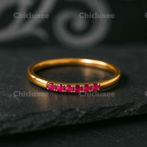 May include: A gold ring with a row of small, vibrant red gemstones. The ring is set against a dark background, highlighting the gold band and the bright red stones. The ring is a piece of jewelry.