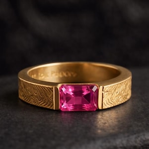 May include: A gold ring with a rectangular, bright pink gemstone. The band has a textured, swirling pattern. The ring is set against a dark background, highlighting the gold and pink colors.
