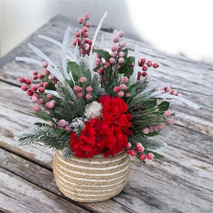 May include: A festive floral arrangement in a woven basket. The arrangement features vibrant red flowers, frosted berries, and evergreen sprigs. Decorative silver accents and white accents add a touch of elegance to this holiday-themed centerpiece.