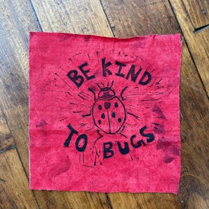 May include: A square red fabric patch featuring a black screen-printed design of a ladybug in the center. The text "BE KIND TO BUGS" is printed in black capital letters in a circular arrangement around the ladybug illustration.