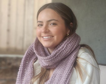 Puede incluir: Una mujer con un suéter blanco y una larga bufanda de punto color malva alrededor del cuello.
