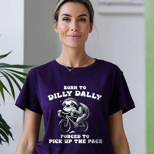 Op de afbeelding: Paars T-shirt met een witte afbeelding van een luiaard die op een fiets rijdt. De tekst op het shirt luidt "Born to Dilly Dally" en "Forced to Pick Up the Pace".