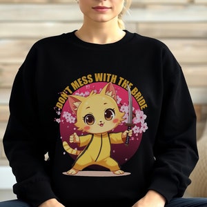 Może przedstawiać: Czarna bluza z nadrukiem kreskówkowego kota w żółtym kombinezonie trzymającego miecz. Kot otoczony jest różowymi kwiatami wiśni i różowym kółkiem z napisem "Nie zadzieraj z panną młodą".