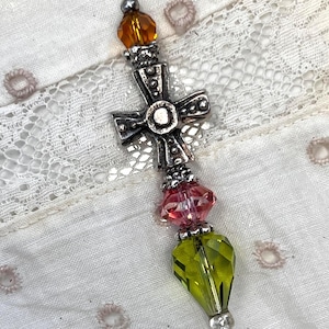 May include: A decorative pin with a silver cross, orange and pink beads, and a green faceted bead. The pin is set against a white fabric background with lace and embroidered details.