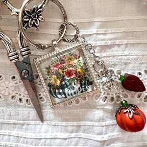 May include: Silver-toned scissors with decorative handles and a square charm featuring a floral arrangement in a pumpkin-shaped container with a black and white checkered pattern. A charm with a pumpkin and leaf completes the design.