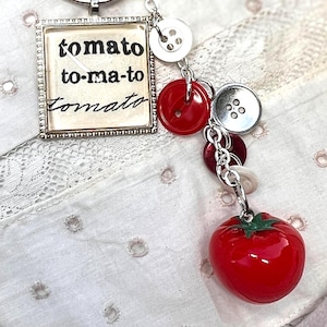 May include: A silver keychain with a square pendant that reads "tomato to-ma-to tomato." The keychain features a charm of red buttons, a red tomato, and white and silver buttons. The tomato charm has a green stem.