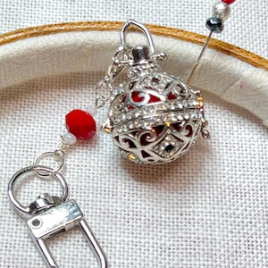 May include: A silver-colored, ornate pendant with a red interior and clear crystal accents. A matching keychain with a lobster clasp is attached. A needle with red and silver beads is also present, suggesting a craft or jewelry-making theme.