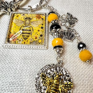 May include: A silver keychain featuring a square pendant with a yellow bee illustration, a round textured charm with a gold bee, and a dangling chain with yellow and black beads, a small silver bee charm, and a miniature silver crown charm.
