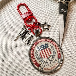 May include: A patriotic keychain with a red clasp and rings, featuring charms of an American flag, a star, and a round pendant. The pendant displays a shield design with the text "America 250 Semiquincentennial 1776-2026 Vintage Stamp USA."