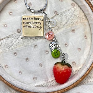 May include: A silver keychain with a square charm featuring the word "strawberry" in a vintage font. The keychain includes decorative buttons and a red strawberry charm. The background is a white fabric.