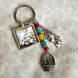 May include: A silver-toned keychain featuring an Easter theme. It includes a square pendant with a bunny and eggs image, colorful beads, and charms of a bunny and a basket. The keychain is designed with a spring holiday aesthetic.