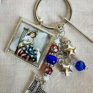May include: A silver keychain with a square pendant featuring a vintage image of a child in patriotic attire. The keychain includes star charms, beads in red, white, and blue, and a small American flag charm.