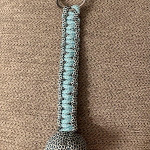 May include: A blue and gray paracord keychain with a knotted ball at the end. The keychain is made of two different colors of paracord, one blue and one gray, and the ball is made of the same paracord.