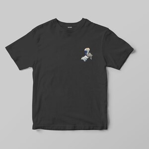 K&ouml;nnte beinhalten: Schwarzes T-Shirt mit einer kleinen, farbenfrohen Grafik auf der oberen linken Brustseite. Die Grafik ist eine stilisierte Illustration mit blauen, wei&szlig;en und grauen Elementen. Das T-Shirt wird vor einem hellgrauen Hintergrund pr&auml;sentiert.