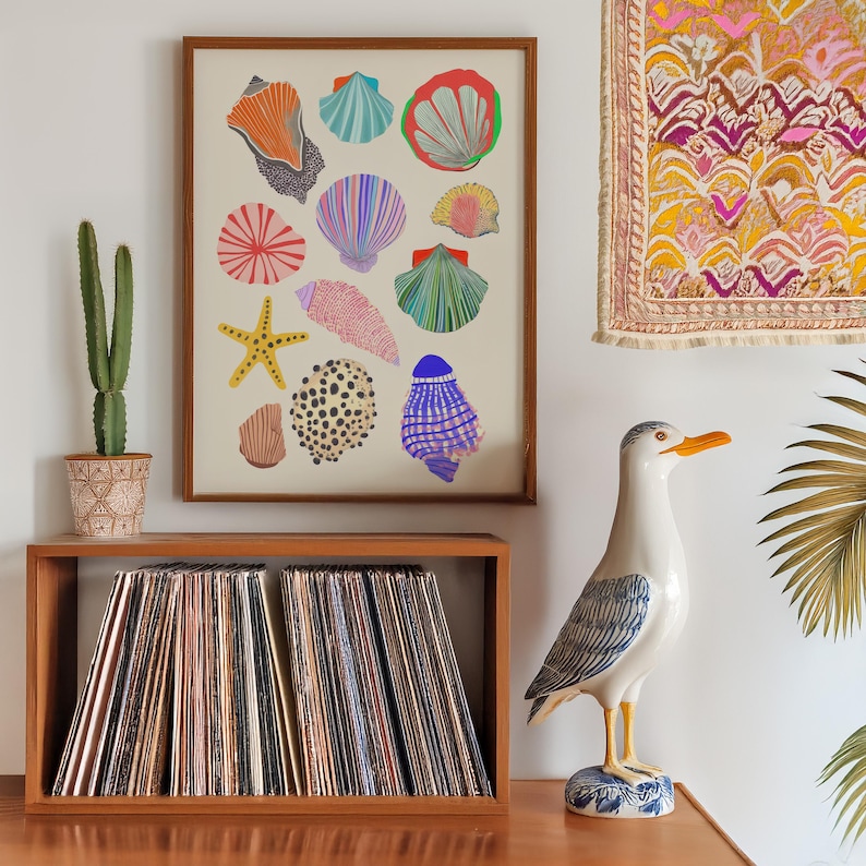 Puede incluir: Una impresi&oacute;n enmarcada con ilustraciones de conchas marinas coloridas cuelga sobre una unidad de almacenamiento de discos de madera. Tambi&eacute;n se ven una estatua de gaviota de cer&aacute;mica y un cactus en maceta. El tema es costero.