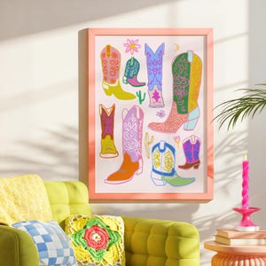 May include: A framed print featuring a colorful illustration of cowboy boots with various patterns and designs. The boots are arranged in a whimsical and eclectic style, with some boots overlapping and others standing alone. The background is a light pink color, and the frame is a light peach color.