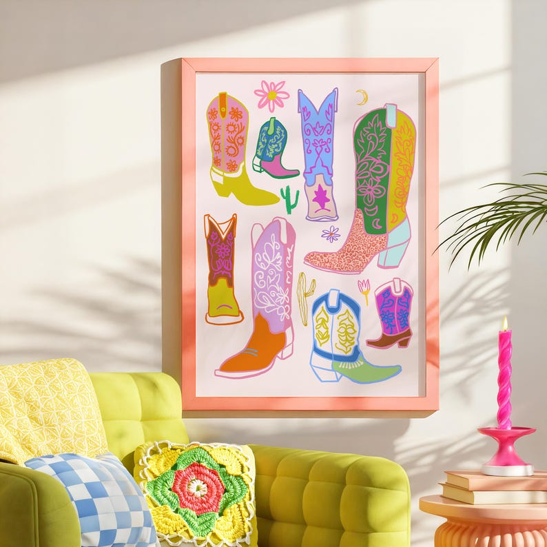 May include: A framed print featuring a colorful illustration of cowboy boots with various patterns and designs. The boots are arranged in a whimsical and eclectic style, with some boots overlapping and others standing alone. The background is a light pink color, and the frame is a light peach color.