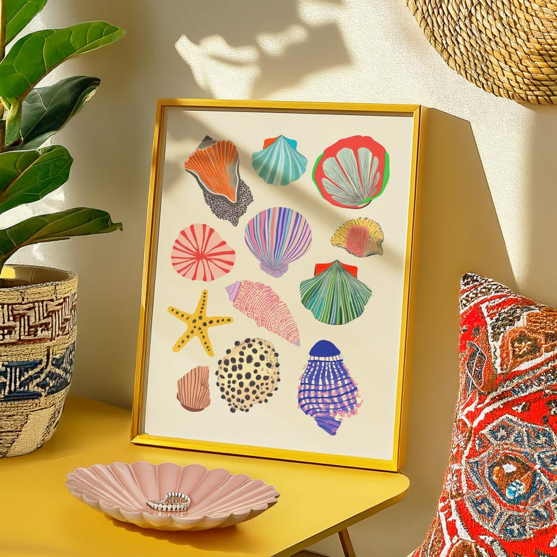 Puede incluir: Una impresi&oacute;n enmarcada con ilustraciones coloridas de conchas marinas y una estrella de mar. La obra de arte est&aacute; en un marco dorado sobre una mesa amarilla. Un plato rosa con volantes y un anillo se encuentra frente a la obra.