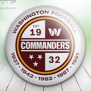May include: A white button with a maroon and gold circular design. The circle has the text "WASHINGTON FOOTBALL" around the outside, "EST 19" and "W" at the top, "COMMANDERS" in the centre, and "32" at the bottom. The text "1937. 1942. 1982. 1987. 1991" is around the outside of the circle.