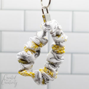 May include: A white fabric key chain with a yellow and gray floral pattern. The key chain is made of a stretchy fabric and has a silver key ring.