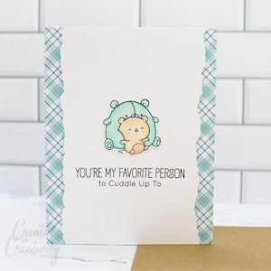 May include: A white card with a green and blue plaid border. The card features a cartoon illustration of two bears hugging. The text on the card reads "You're my favorite person to cuddle up to."