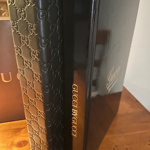 May include: Two black leather-bound books with gold lettering. The book on the left has a repeating Gucci pattern on the cover. The book on the right has the text "Gucci by Gucci" on the spine and "Gucci" on the front cover.