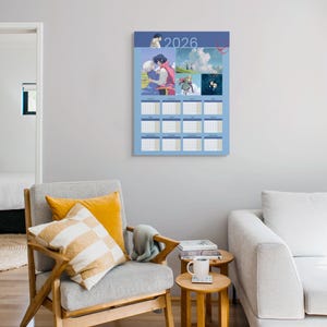 May include: A blue 2026 calendar featuring anime-style illustrations. The calendar is hanging on a gray wall above a gray armchair with yellow pillows. A small wooden side table with a mug and books is next to the chair.