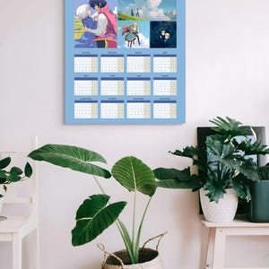 May include: A light blue 2026 calendar featuring anime-style illustrations. The calendar has a grid layout with dates and months. The top section includes several colorful images. The calendar is hanging on a white wall, with plants in the foreground.