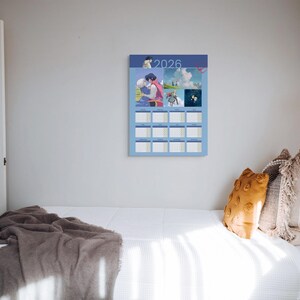 May include: A blue 2026 calendar with anime-style illustrations hangs on a white wall. The calendar has a grid layout with monthly boxes. A bed with white bedding and decorative pillows is in the foreground.