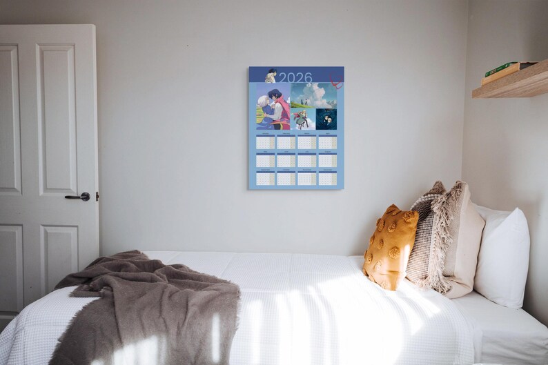 May include: A blue 2026 calendar with anime-style illustrations hangs on a white wall. The calendar has a grid layout with monthly boxes. A bed with white bedding and decorative pillows is in the foreground.