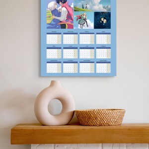 May include: A 2026 calendar featuring anime-style illustrations, with a light blue background and monthly date grids. The calendar is displayed on a wall above a wooden shelf with a beige vase and a woven basket.