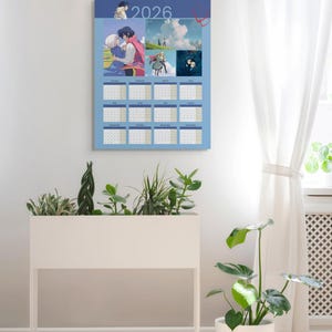 May include: A 2026 calendar with a light blue background, featuring anime-style illustrations. The calendar has a grid layout with each month displayed. The image is hanging on a white wall, with a white planter box and a potted plant in the foreground.