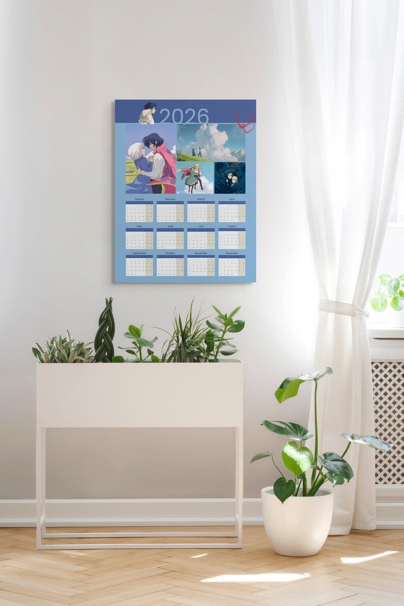 May include: A 2026 calendar with a light blue background, featuring anime-style illustrations. The calendar has a grid layout with each month displayed. The image is hanging on a white wall, with a white planter box and a potted plant in the foreground.