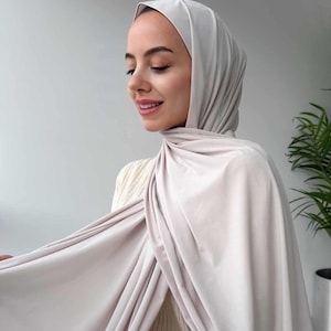 Op de afbeelding: Een lichtgrijze hijab, elegant gedrapeerd. De stof lijkt zacht en vloeiend, met subtiele plooien en texturen. De hoofdbedekking wordt door een persoon gedragen, met een neutrale achtergrond en een glimp van een groene plant.