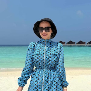 Puede incluir: Una persona con un vestido azul estampado con un sombrero negro y gafas de sol está en una playa. El vestido tiene un diseño geométrico en negro y azul, una cremallera y mangas largas. El fondo presenta un océano turquesa y un cielo azul claro.
