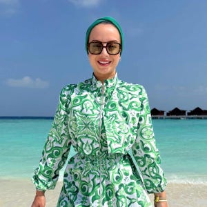 Puede incluir: Una persona con un vestido estampado en verde y blanco con un pañuelo en la cabeza verde a juego y gafas de sol. El vestido tiene cierre de cremallera y mangas largas. El fondo muestra una playa con agua turquesa y cielo azul claro.